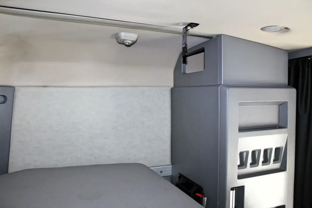 2023 Mack Anthem — sleeper lower bunk mattress, cream padded wall, ceiling dome light, gray cabinet with vents and dark curtain
