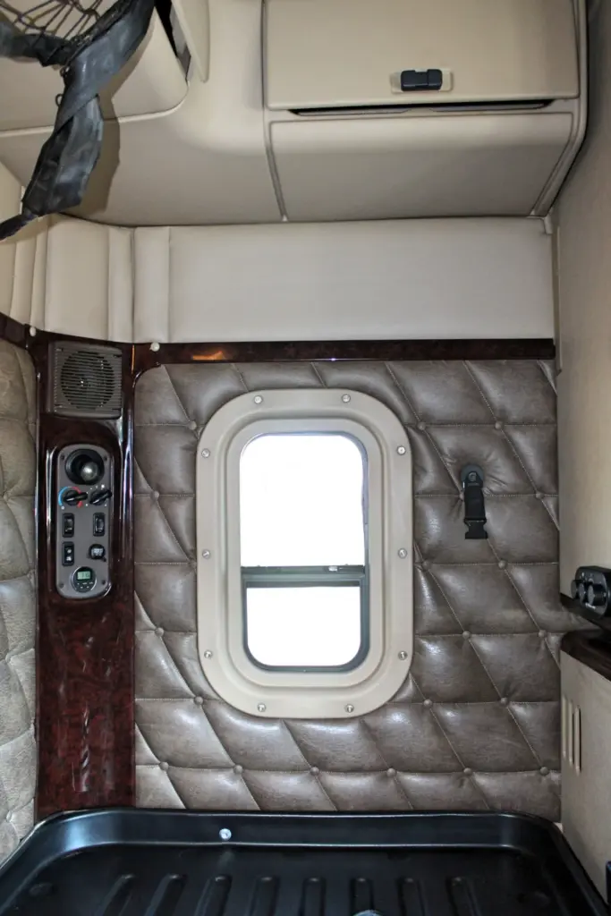2016 Freightliner Coronado 132 — sleeper wall with rounded window, quilted brown panel and wood-grain controls by a speaker
