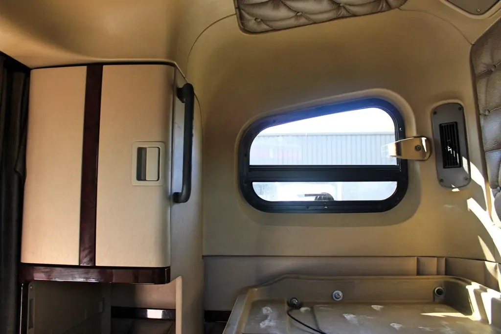 2020 Freightliner Coronado 132 — wide view of tan sleeper wall with a rectangular window, wood-trim cabinet and wall vent