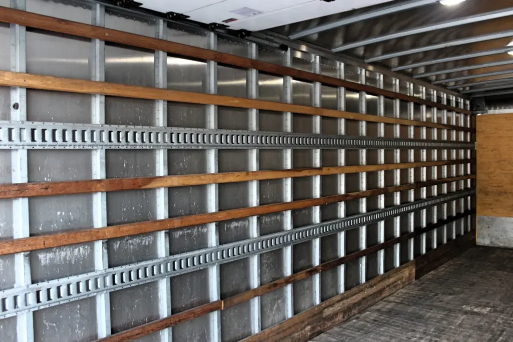 2020 Freightliner M2 106 — angled view inside empty box body, wood-slatted walls, metal tie rails and plywood front wall