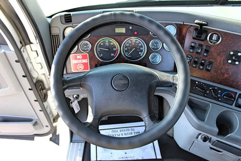 2020 Freightliner Columbia 120 — driver's-seat view of black leather-wrapped wheel, wood-grain dash and round gauges.