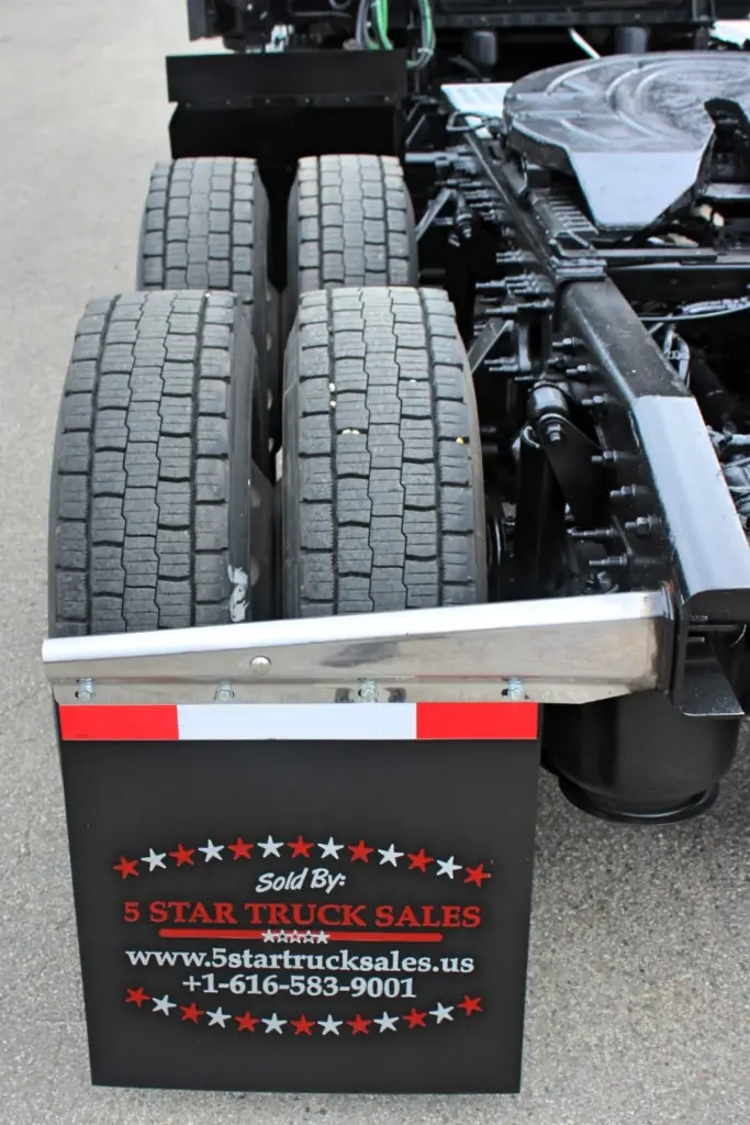 2021 Freightliner Cascadia 126 — low close-up of tandem tires with steel-trimmed mud flap and black air tanks beneath frame.